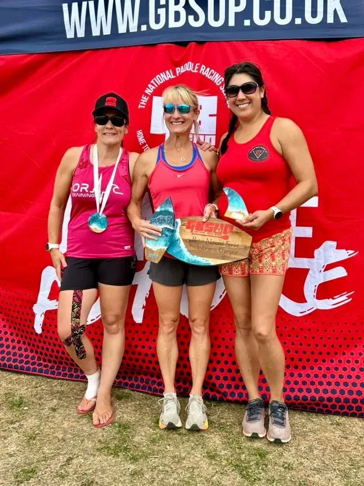 Three women stand together in front of a red banner. The woman in the middle holds an award, the others hold medals, smiles on their faces.