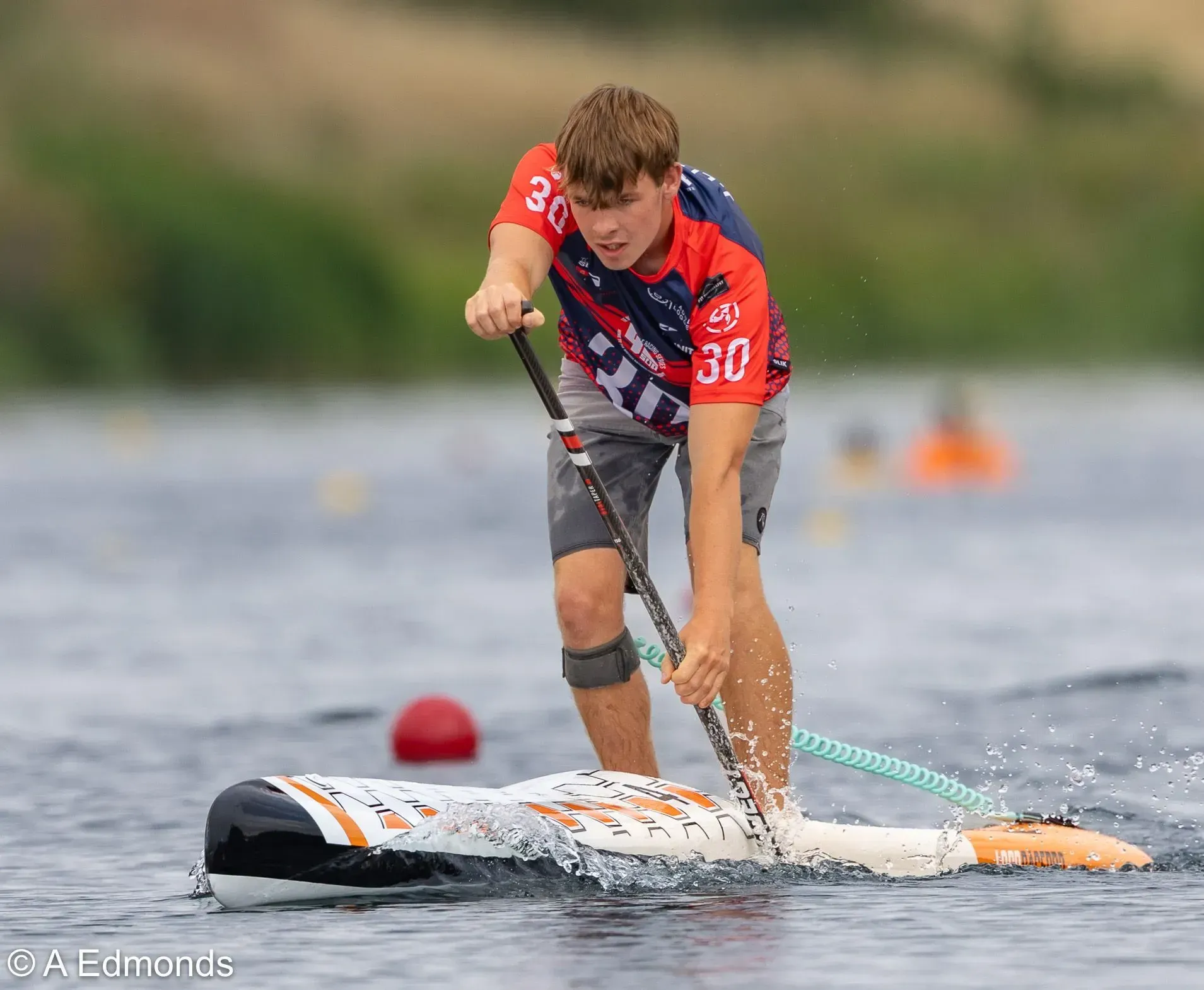 A young man in a red jersey races a stand-up paddleboard on a lake. He leans forward, pulling the paddle through the water.