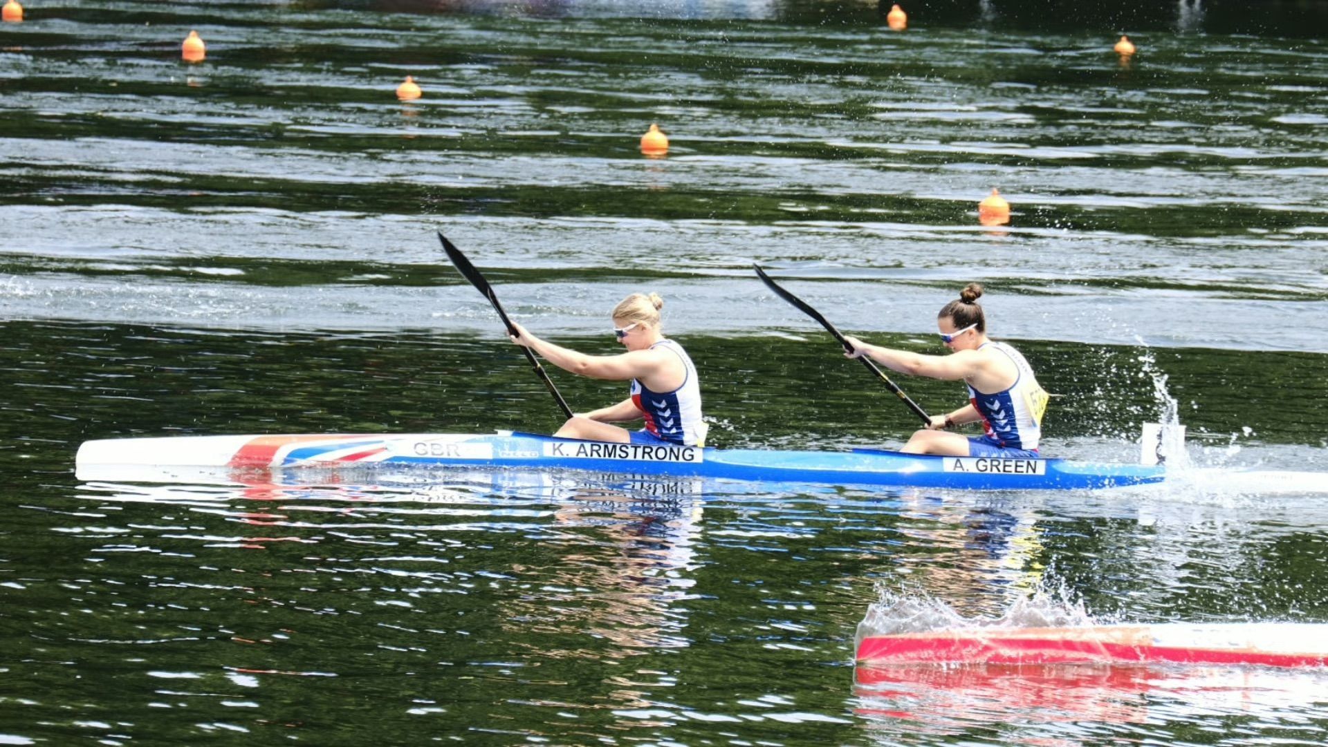 Two female athletes in a kayak rowing on a lake. They wear blue and white uniforms, rowing in unison.