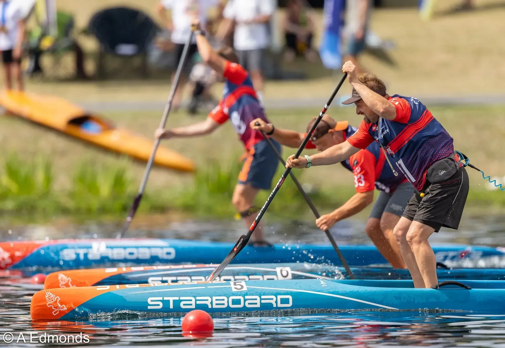 Three paddlers in a Stand Up Paddleboard race on a lake, paddling hard. Wearing matching red, blue, and white uniforms;