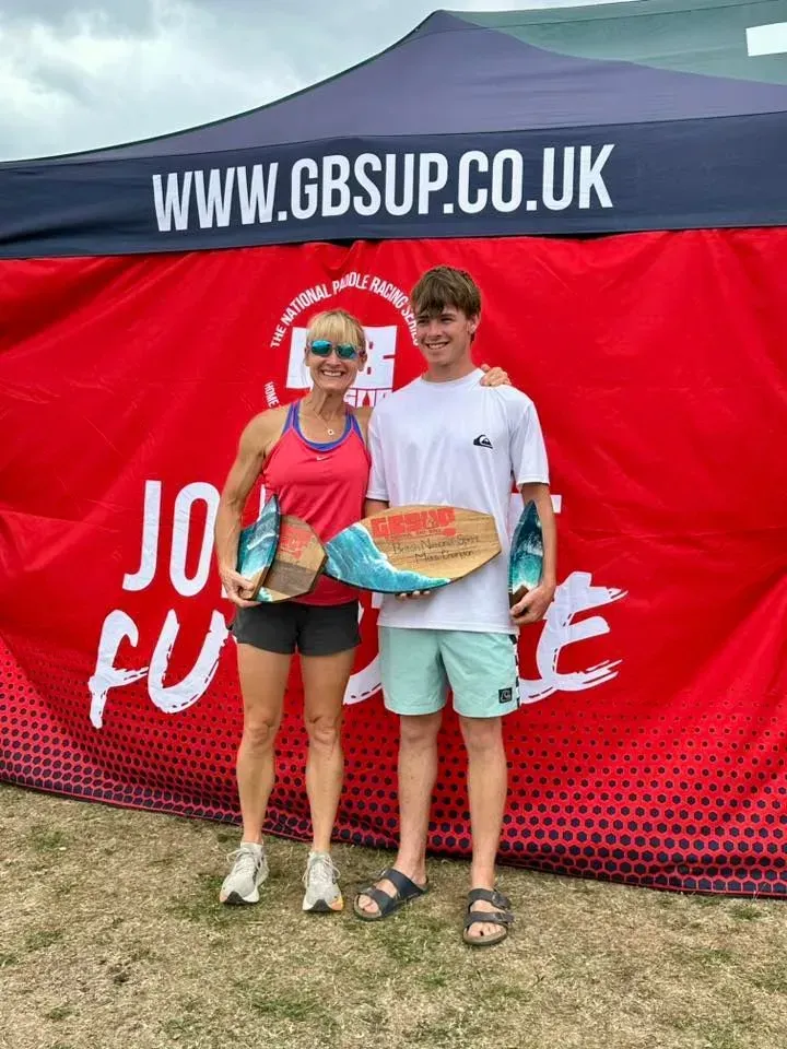 A woman and a teen boy smiling, holding awards, standing in front of a red and blue banner. They're outside on a grassy area.