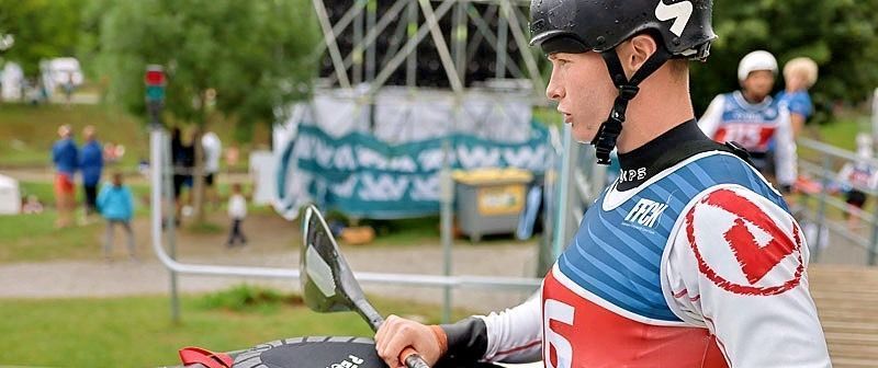 A kayaker in a red and blue uniform holds a paddle, preparing to compete. He wears a helmet and stands near a metal structure.