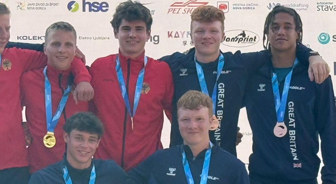A group of young men stand together, wearing medals and athletic apparel, on a podium outdoors. Some are smiling and posing.