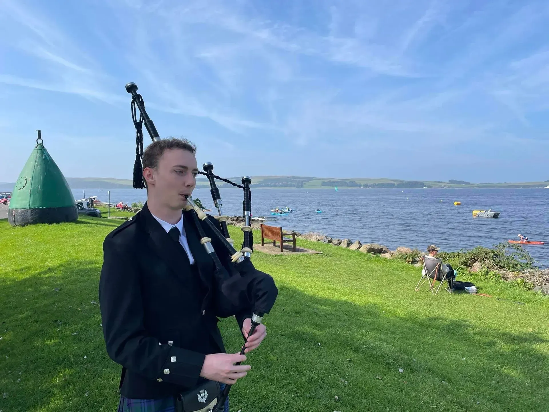 A young man in a kilt plays bagpipes on a grassy bank by a blue lake under a sunny sky.