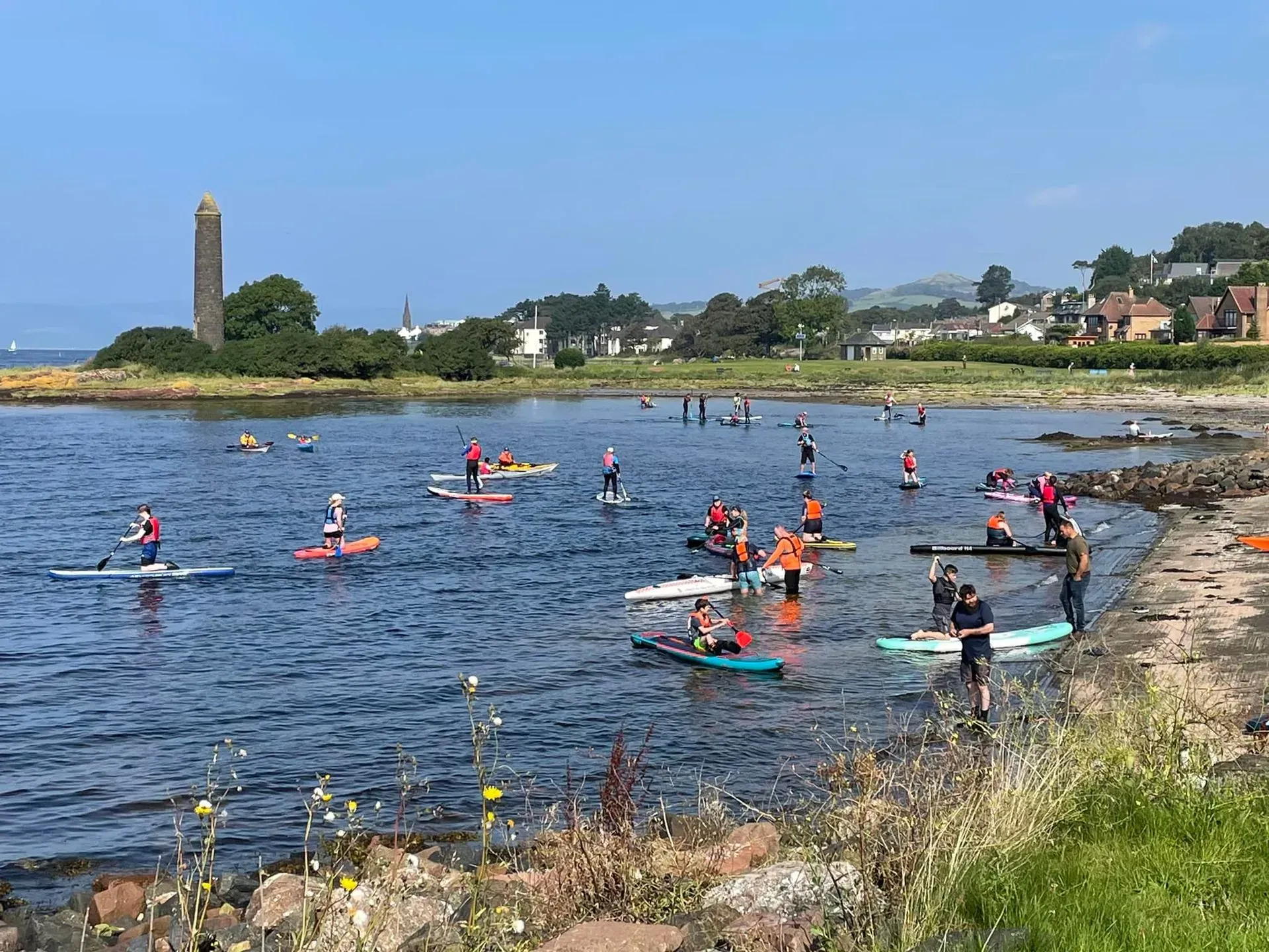 People on paddleboards in a bay near a grassy shore on a sunny day, with a tall stone monument in the background.