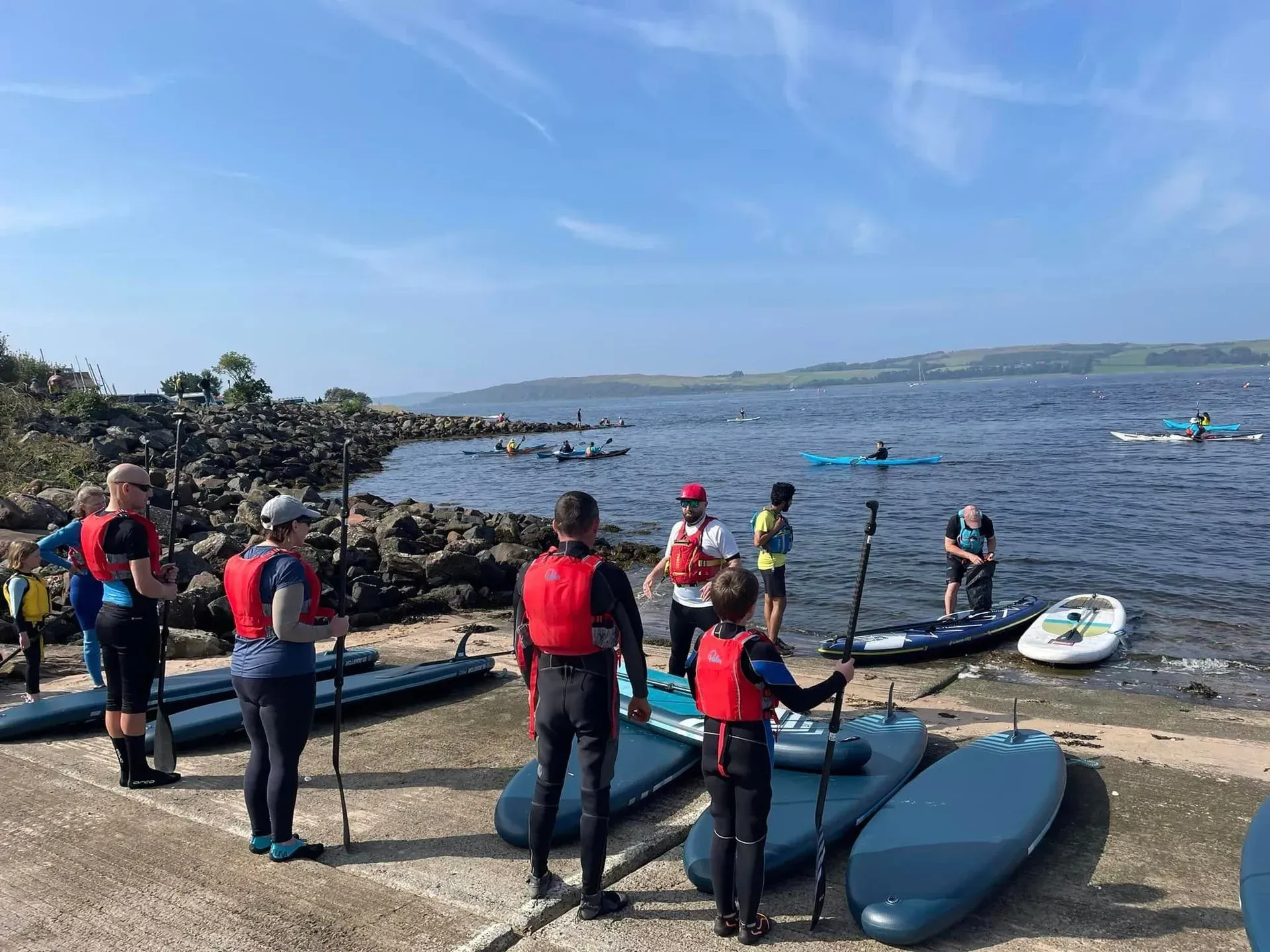 People preparing to paddleboard on a lake. Several paddleboards and individuals wearing life vests stand on a shoreline under a blue sky.