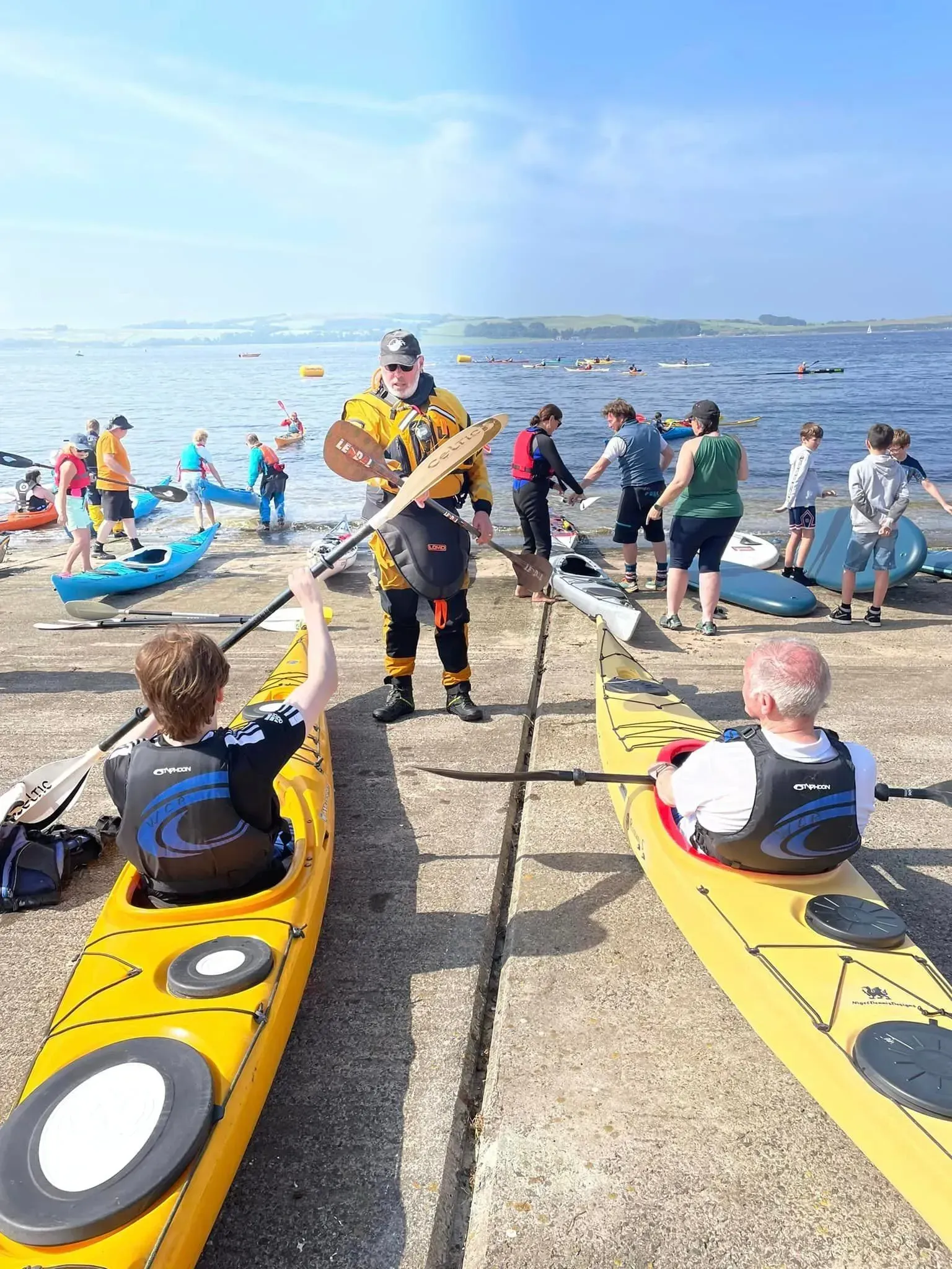 People preparing to kayak on a sunny day. A kayaker in yellow gear instructs two others in yellow kayaks, with other kayakers in the background.