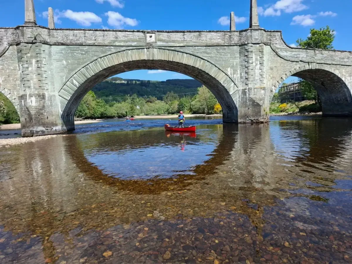 A person is in a red canoe under a bridge over a river.