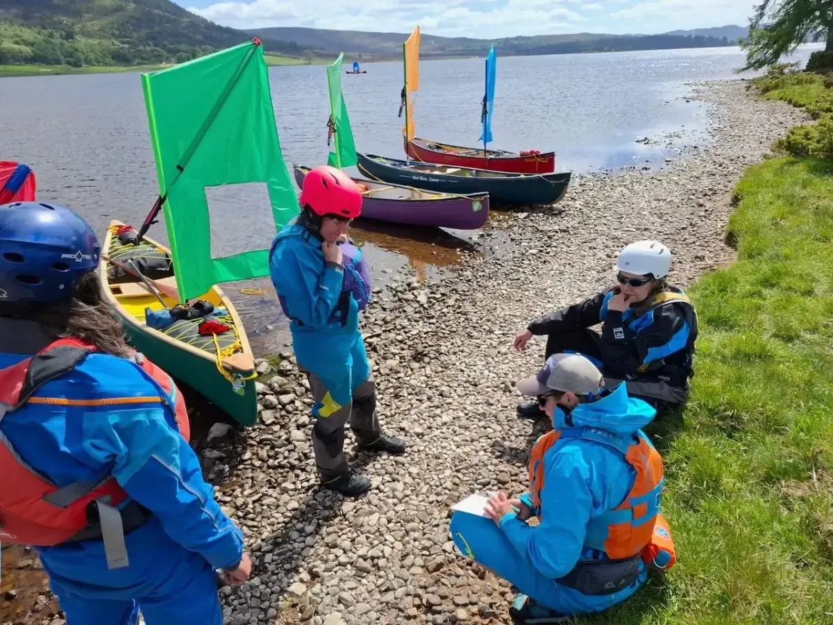 People in wetsuits near colorful sailboats on a pebbled beach by a lake. They appear to be discussing something, with one person taking notes.