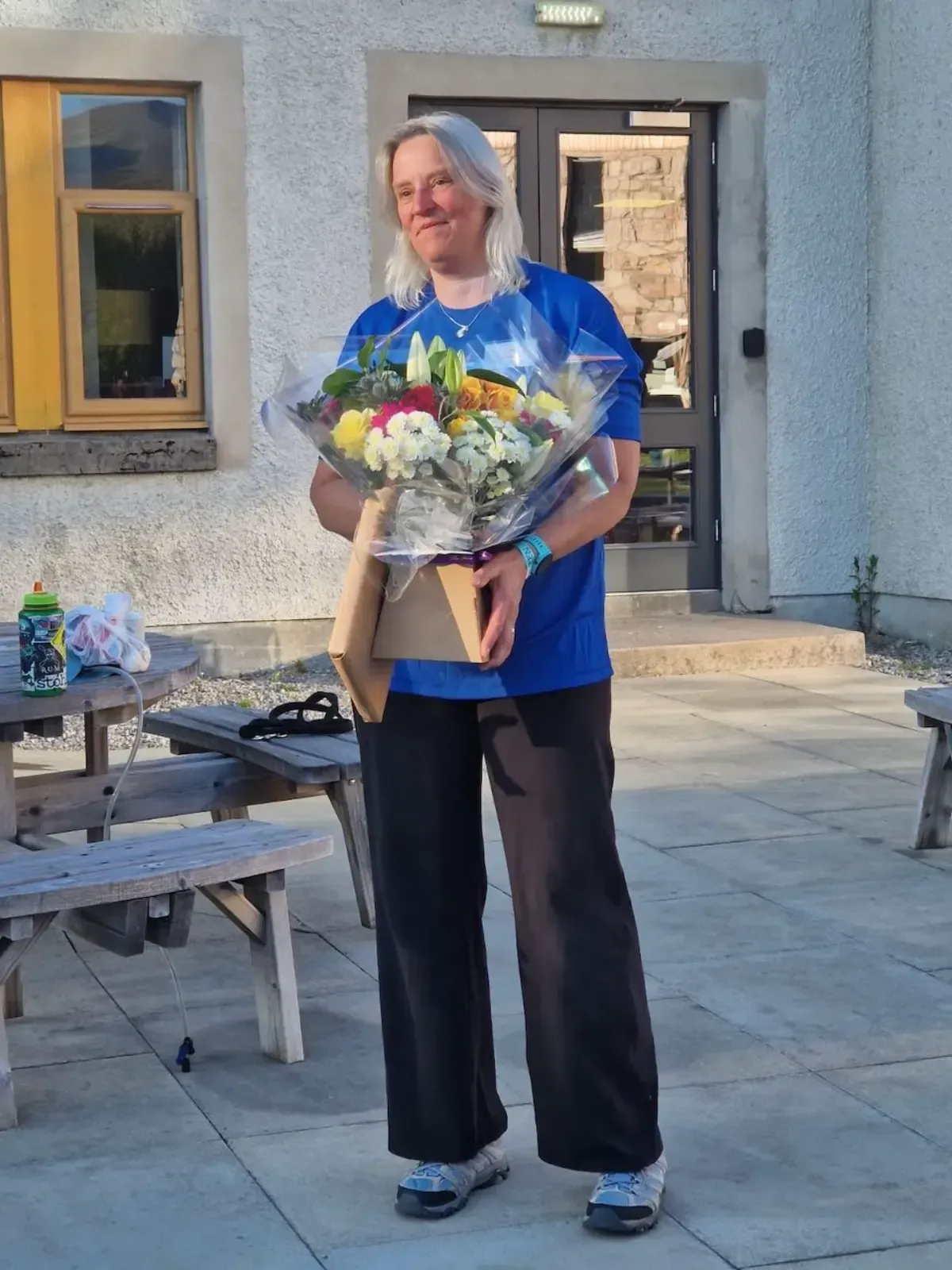 Woman holding a bouquet of flowers, wearing a blue shirt and black pants, standing outside a building.