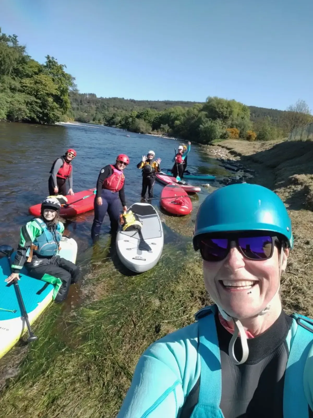 A group of people, wearing safety gear, prepare to paddleboard and kayak on a river on a sunny day. A woman in the foreground smiles for a selfie.