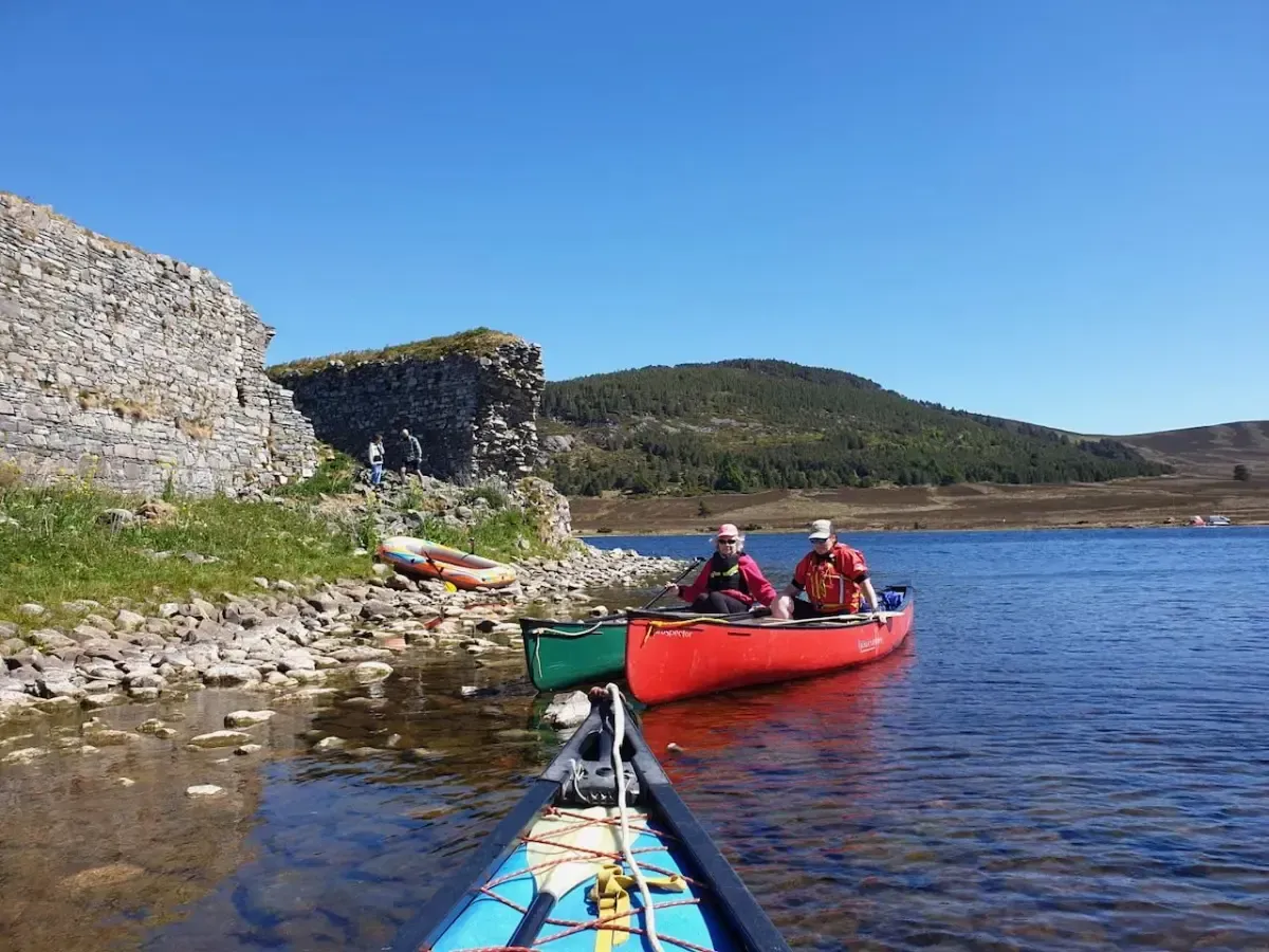 Two people canoeing on a lake, near a stone wall and a mountain. Clear blue sky.