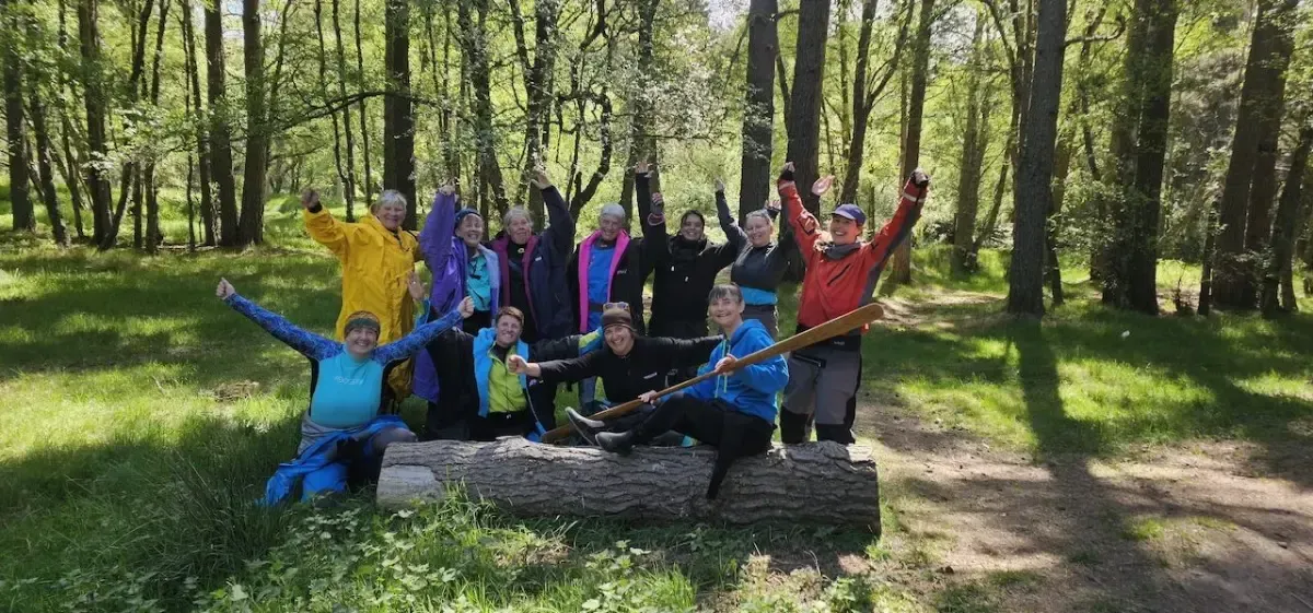A group of people in colorful outerwear pose in a forest, some with arms raised, on and around a log.