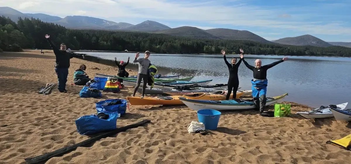 People with arms raised on a sandy beach beside a lake, with kayaks and mountains in the background.