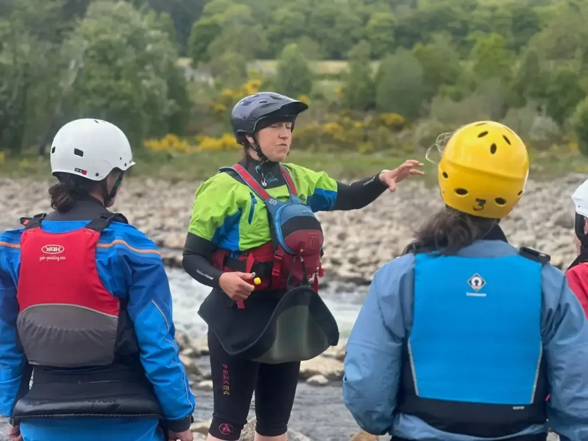 A group of people wearing life jackets and helmets are standing next to a river.