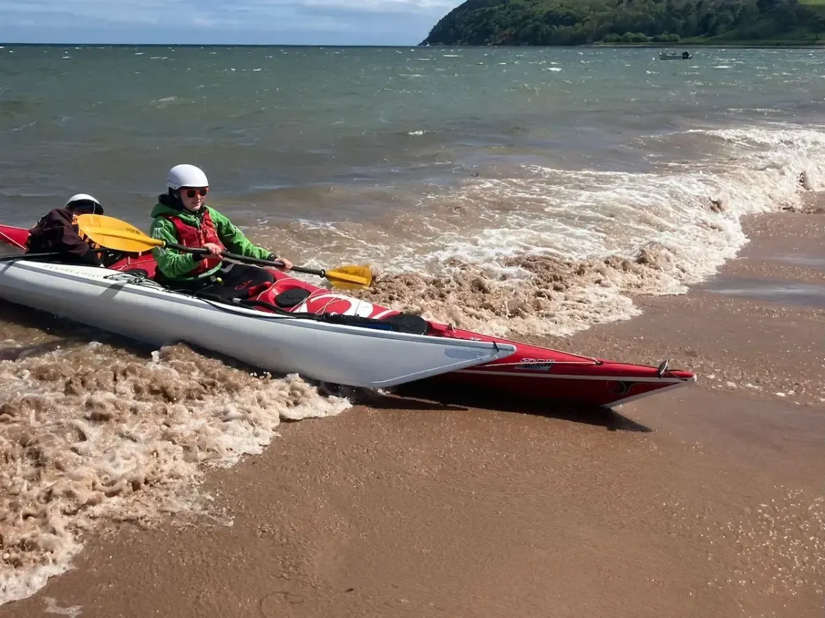 Kayaker in a white and red kayak on a sandy beach, paddling toward shore. They are wearing a helmet and green jacket.