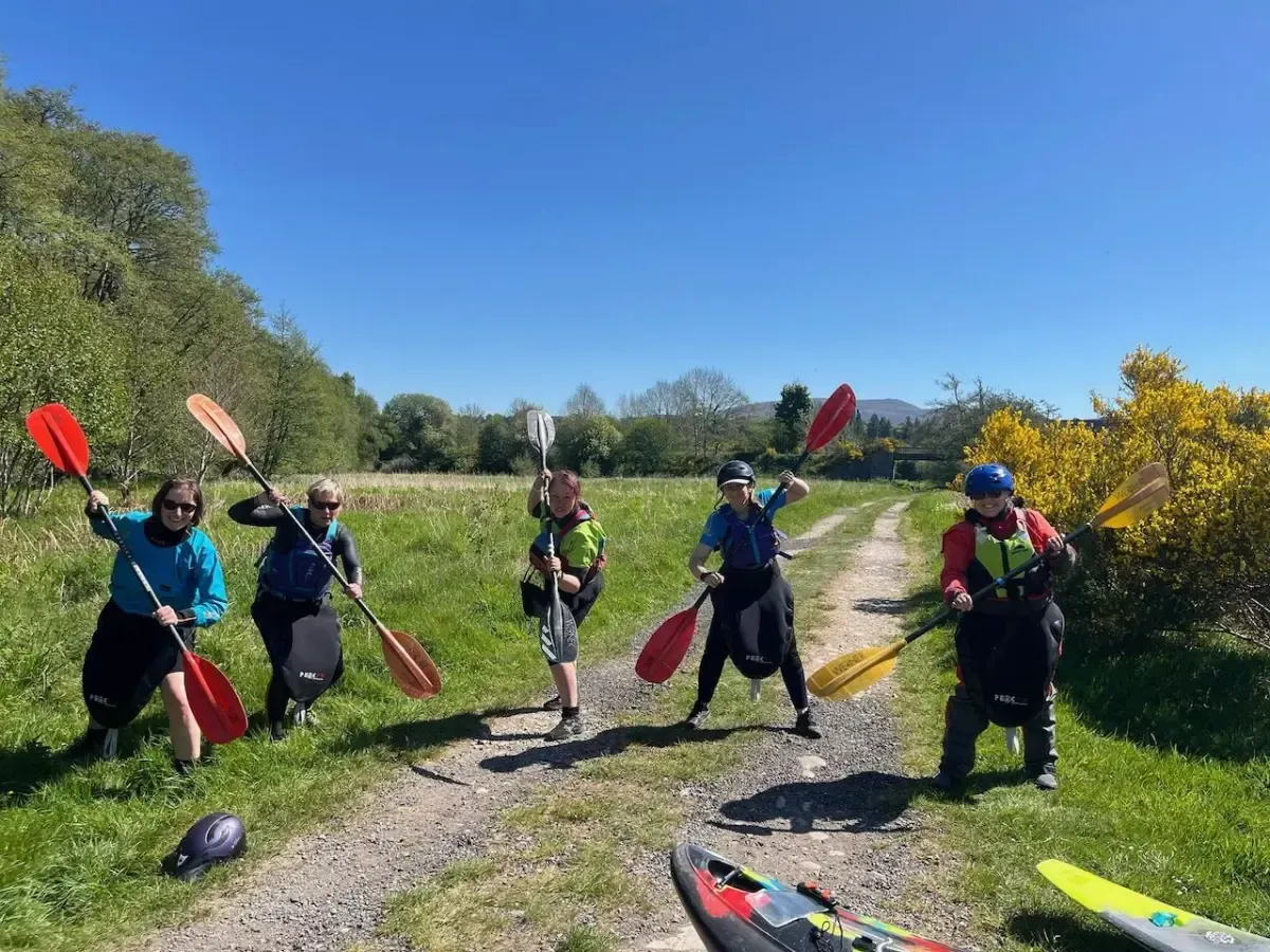 Group of kayakers holding paddles, posing on a grassy path under a blue sky. They're wearing gear like life vests and drysuits.