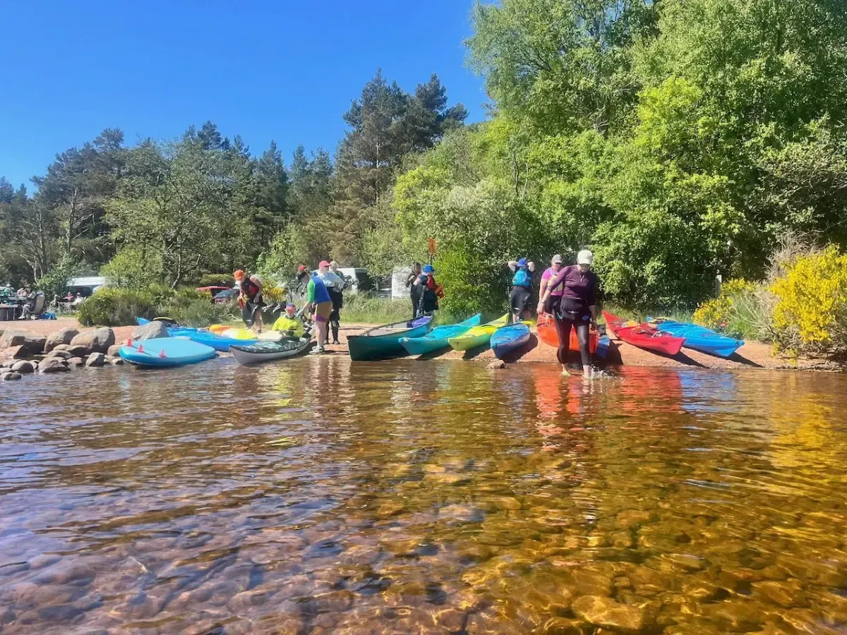 People with kayaks on a shoreline with clear, shallow water.  They are gathered near the boats, with trees and blue sky in the background.