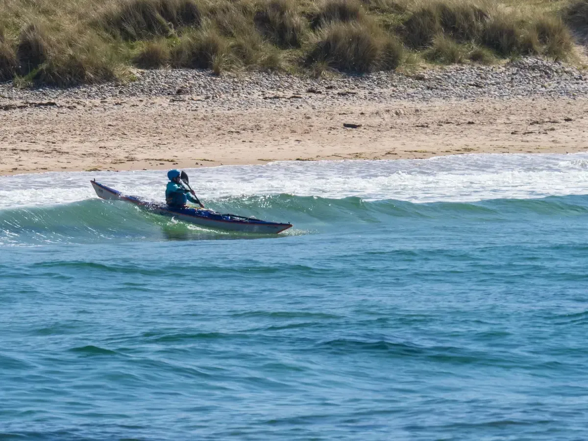 A person in a kayak surfs a small wave near a sandy beach and dunes, blue water and sky.
