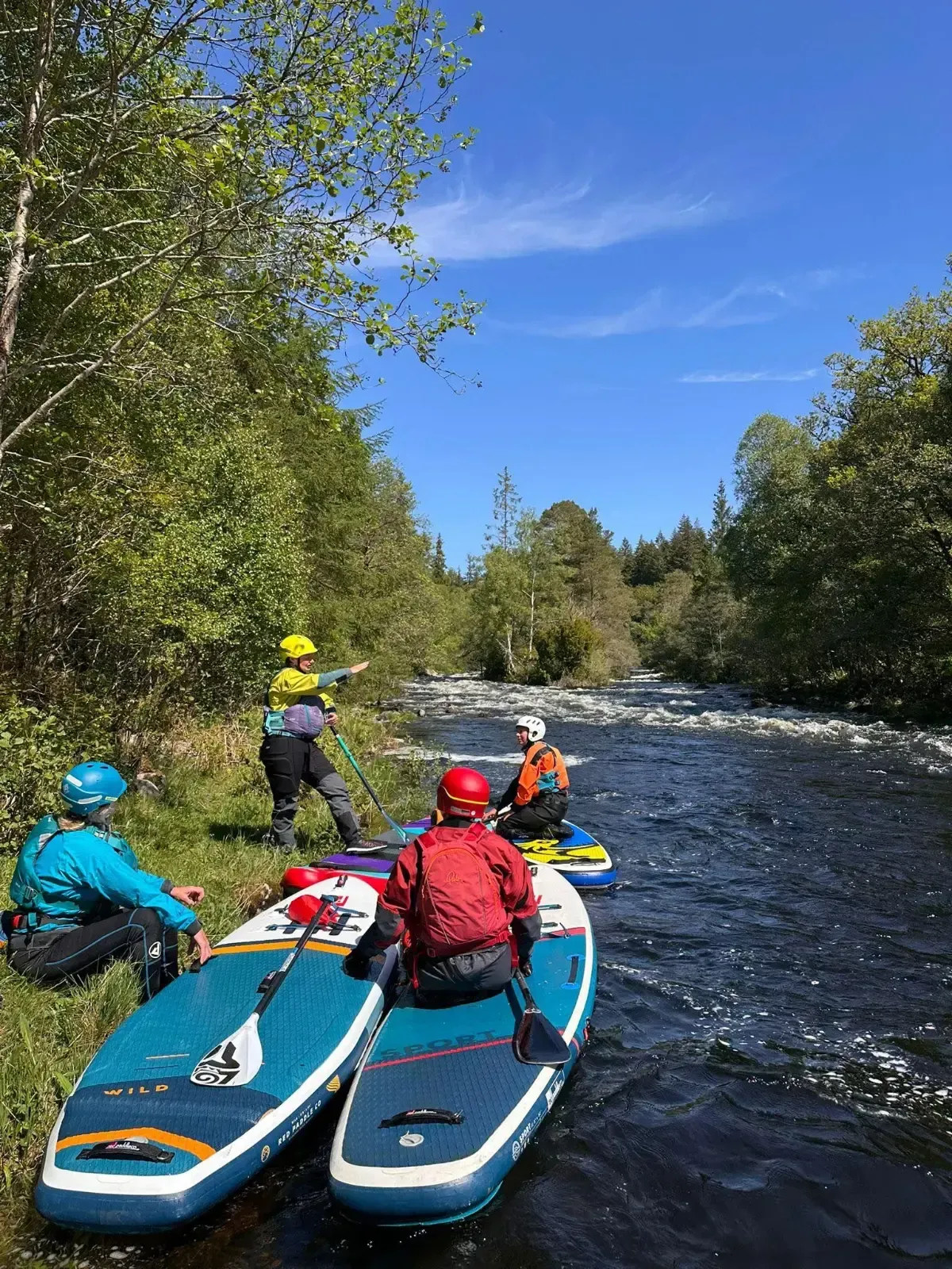 People on paddleboards near a riverbank. They wear wetsuits and helmets. The setting is a sunny day with trees lining the river.
