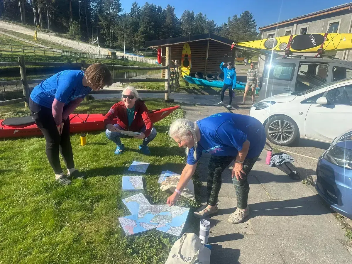 Three women in blue shirts study maps on the ground outdoors near kayaks and a building.