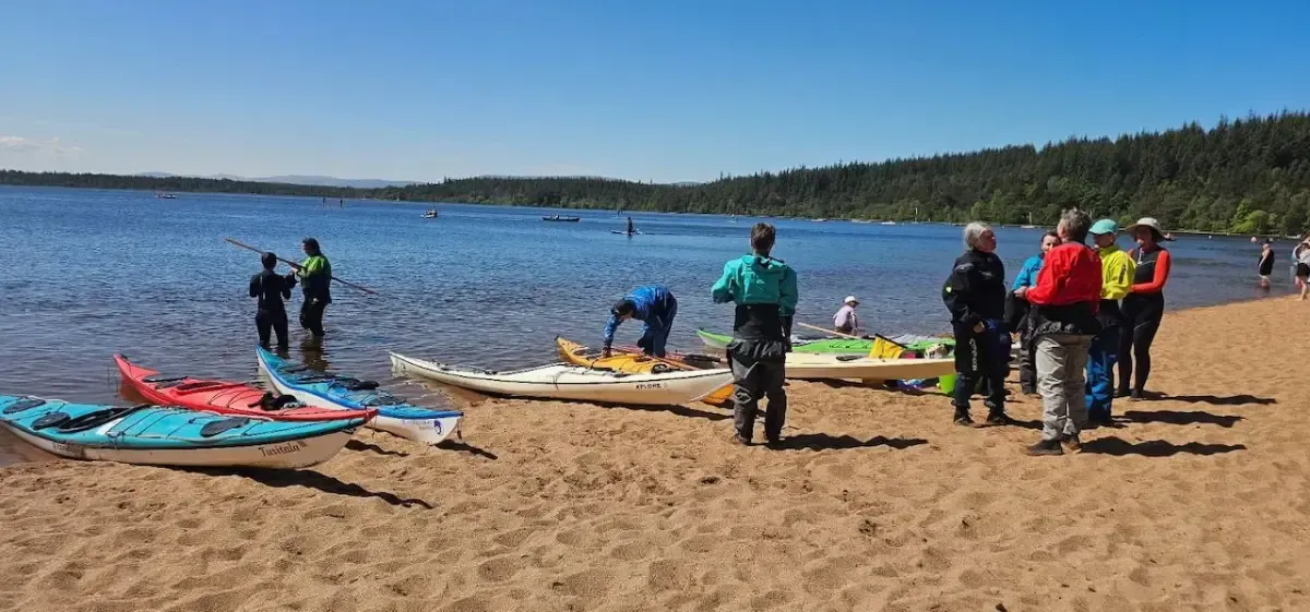 People preparing kayaks on a sandy beach with a lake and trees in the background on a sunny day.