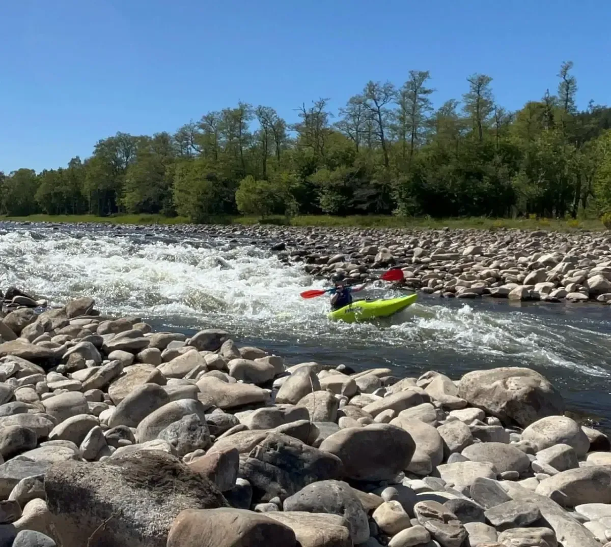 Kayaker in a lime green boat paddles through whitewater rapids on a river surrounded by rocks and trees under a blue sky.