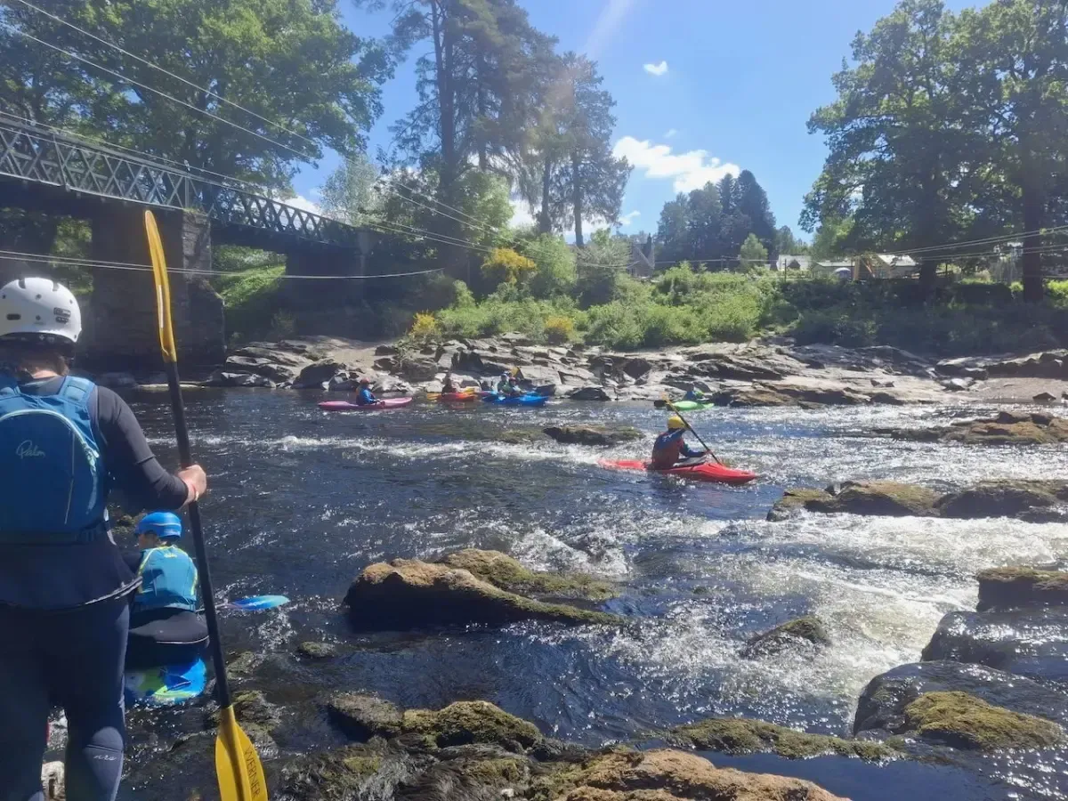 Kayakers navigate a river with rapids under a bridge. Sunlight glints off the water.