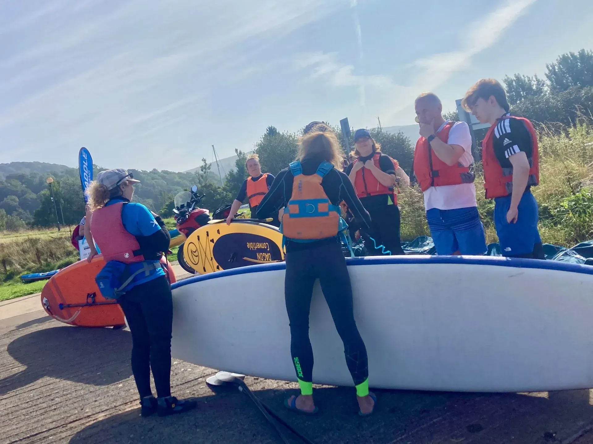 People wearing life vests and wetsuits preparing to go paddleboarding on a sunny day. A paddleboard is lying on the ground.