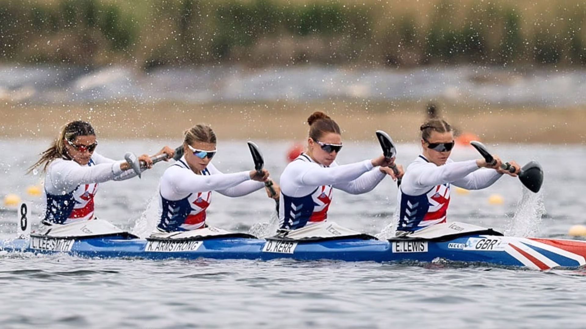 Four women in a blue kayak racing on water. They are paddling in unison, wearing white shirts and sunglasses.