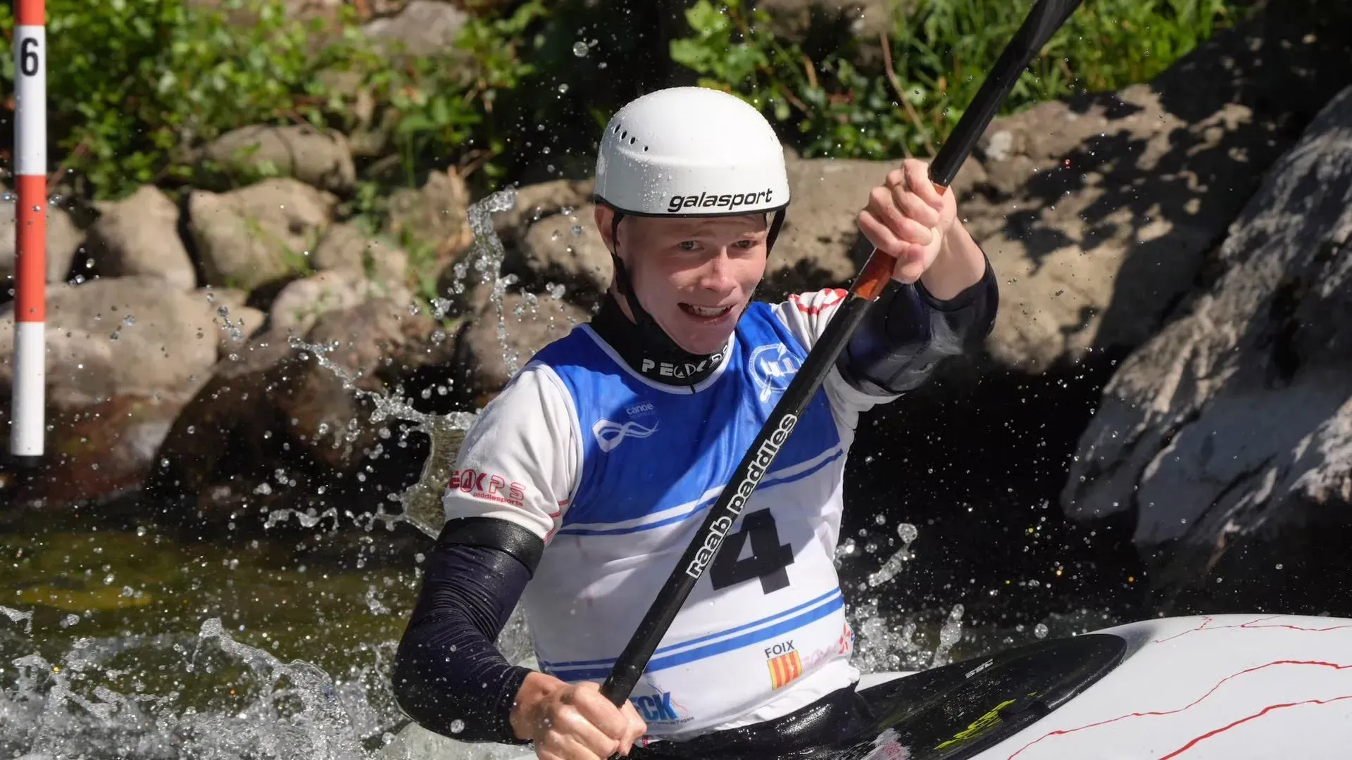A young kayaker navigates a whitewater course, paddling vigorously. They wear a helmet and competition gear in blue, white, and orange.