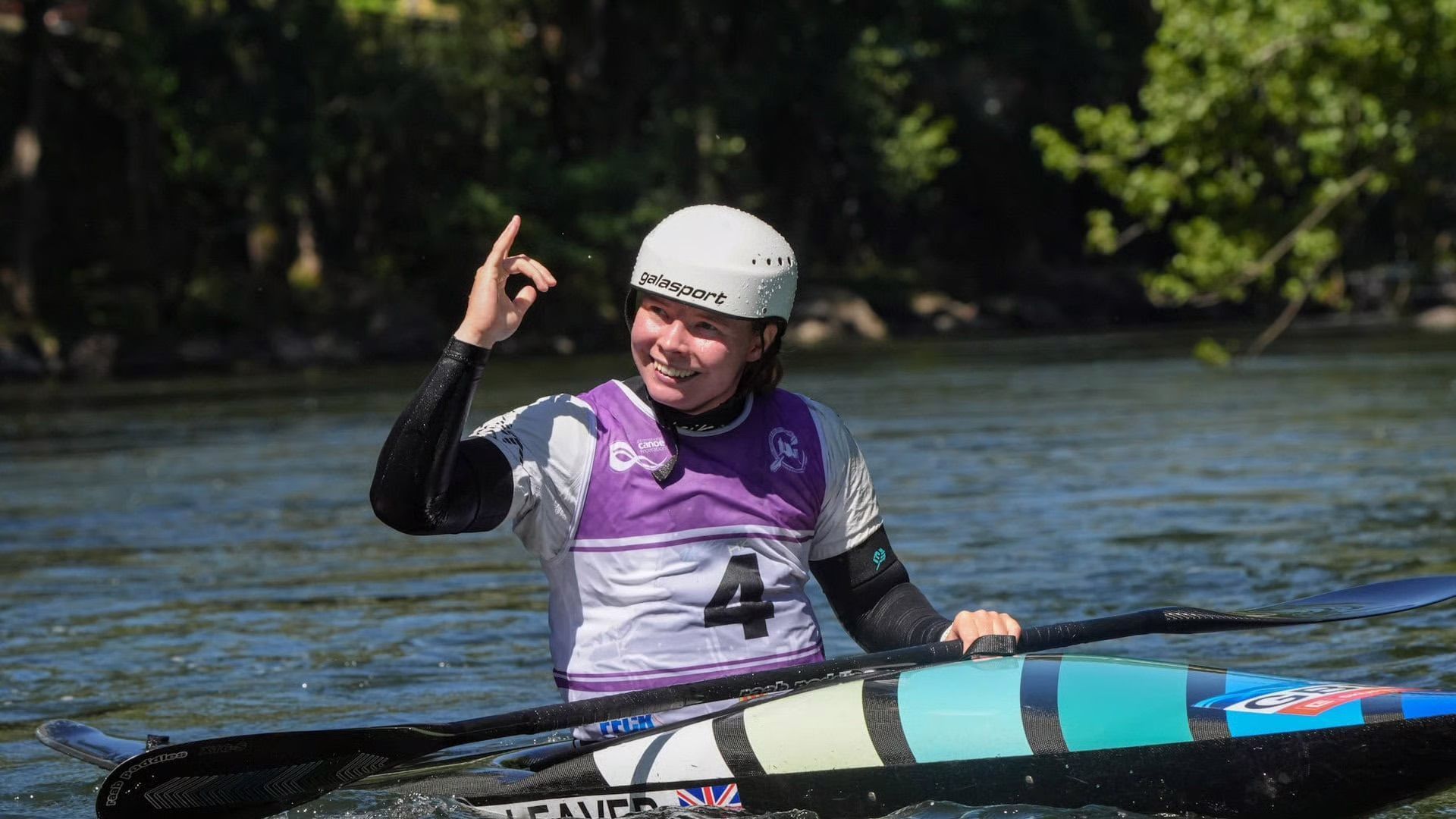 Female kayaker in white helmet and purple jersey raises a finger, smiling on a river.