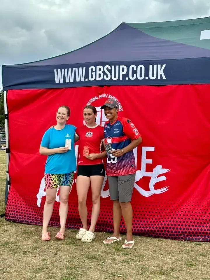 Three people stand in front of a red banner, holding trophies. A woman in a red shirt and shorts is in the center.