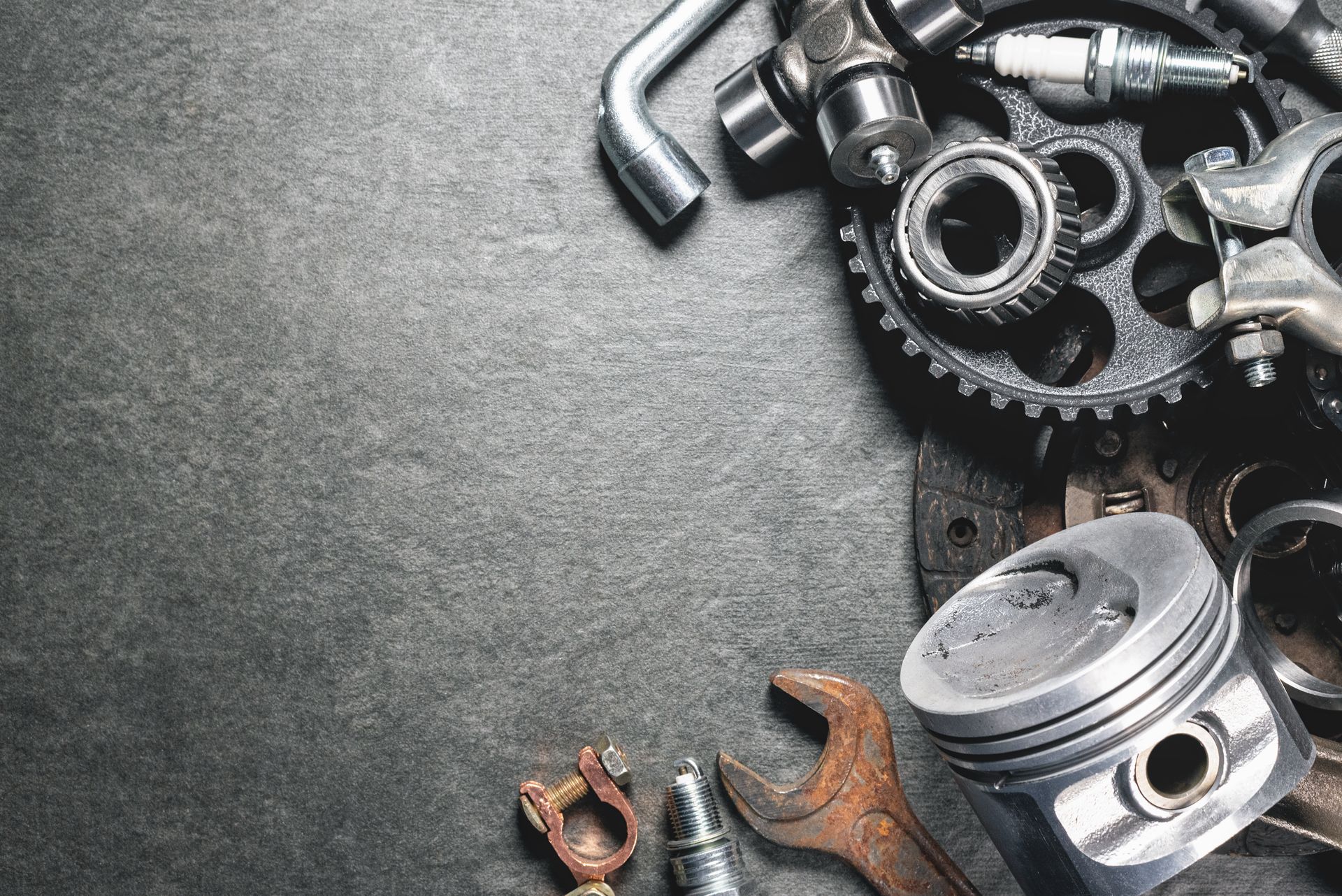 Assorted metal gears, bolts, and tools scattered on a dark textured surface. Automotive repair tools and engine components arranged on a metal workshop surface.
