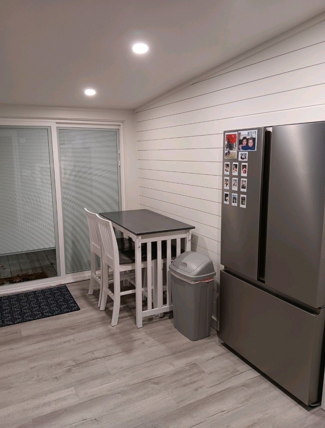 Modern kitchen with stainless fridge, small dining table, white chairs, and sliding glass doors.