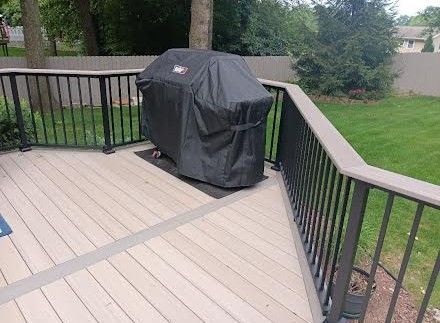 Covered black grill on a wooden deck with black railing, surrounded by trees.