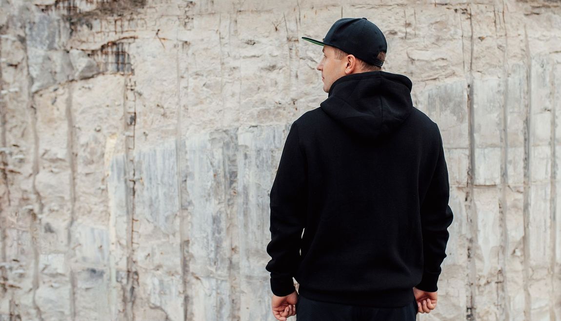 Man in black hoodie and cap, standing in front of a textured concrete wall, looking away.