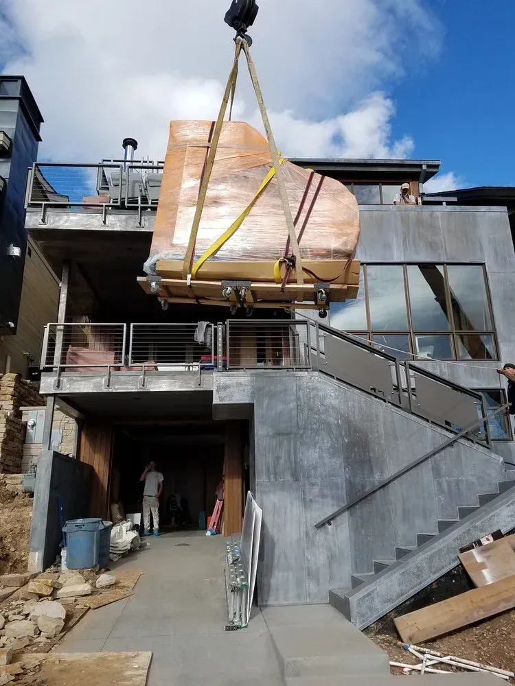 A piano, wrapped for transport, is hoisted by a crane to a house with a concrete exterior.