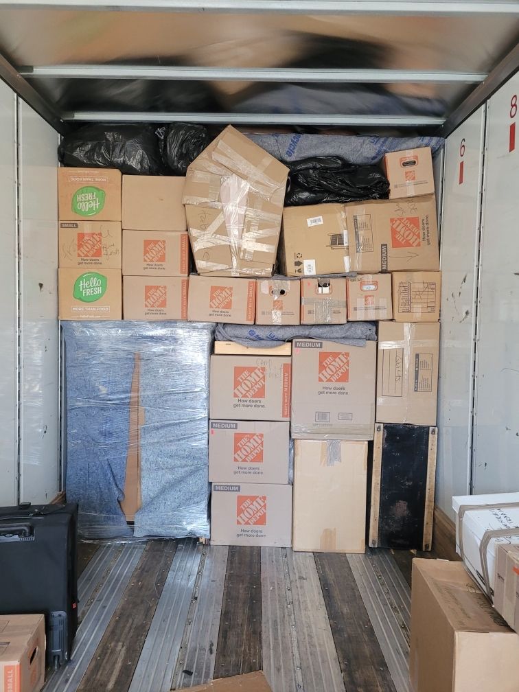 Interior of a moving truck packed with cardboard boxes and furniture wrapped in plastic, ready for transport.