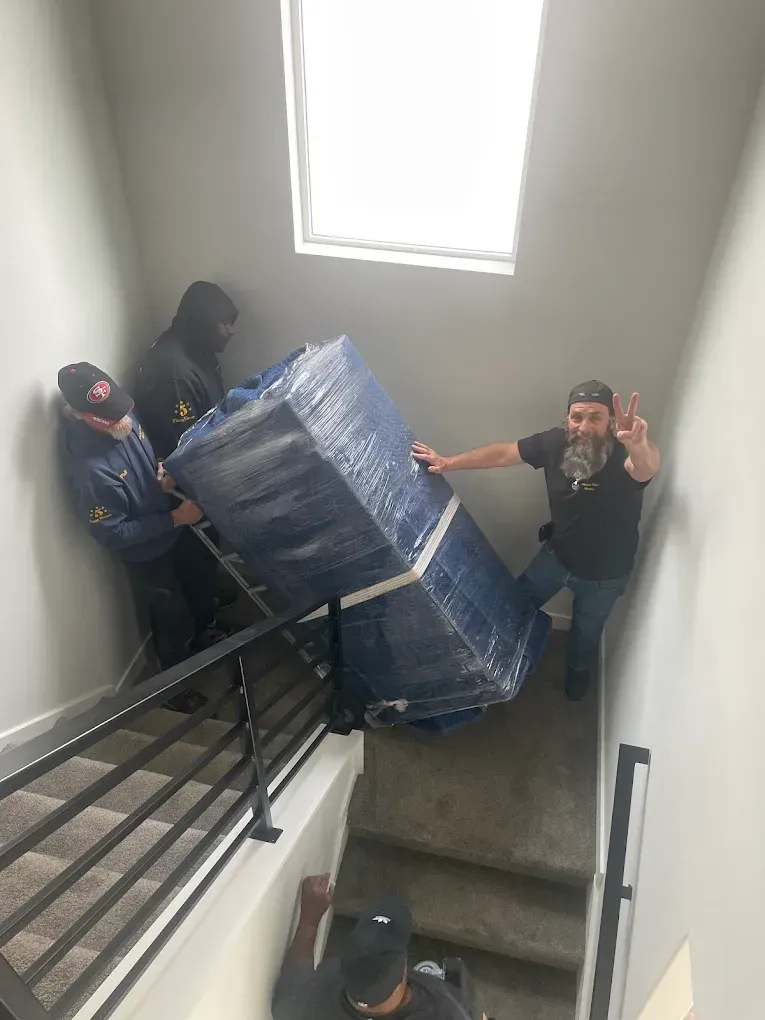 Movers carrying a large, wrapped object up a staircase. One waves.