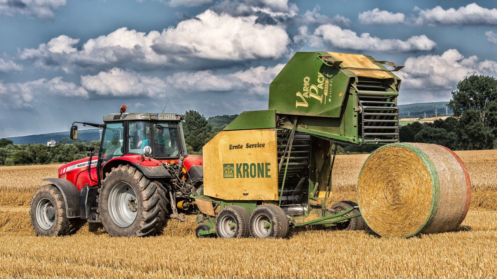 Red tractor towing a green John Deere baler in a wheat field with round hay bales and cloudy sky