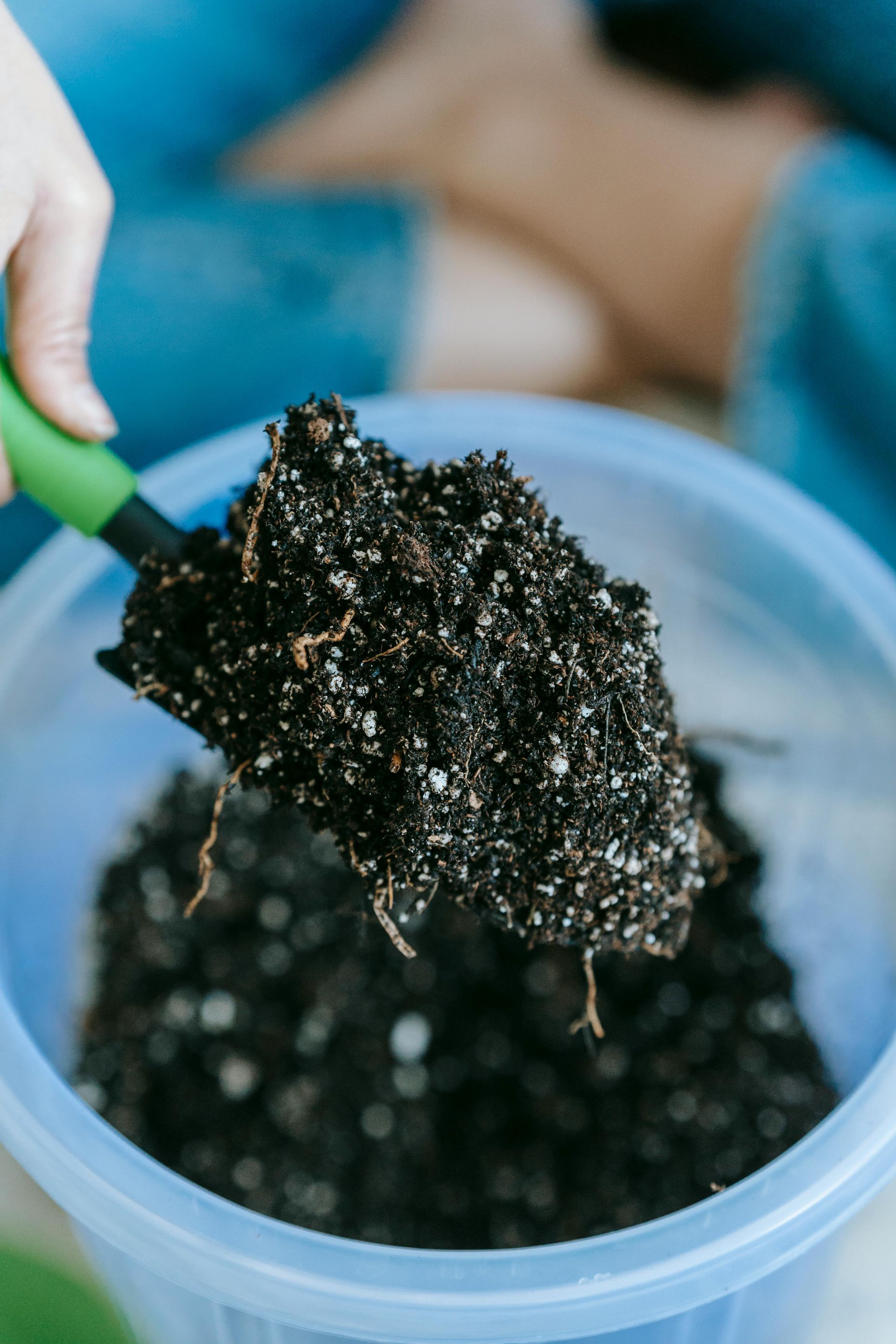 Person using a green-handled trowel to scoop soil from a blue bucket.