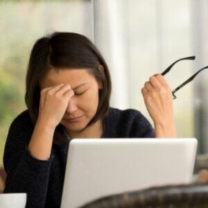 A woman is sitting in front of a laptop computer holding her glasses.