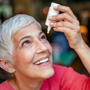 A woman is smiling while holding a bottle of eye drops in her hand.