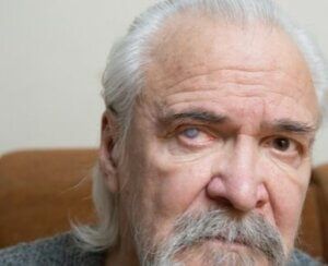 An older man with a beard and gray hair is sitting on a couch.