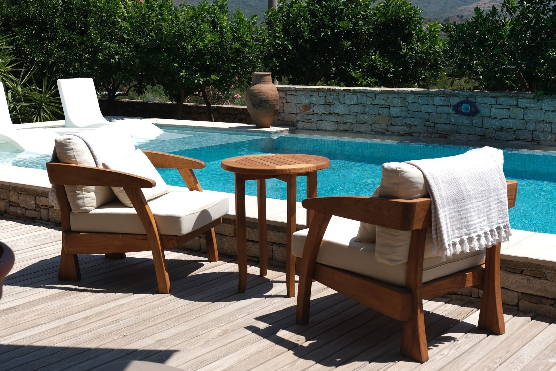Two white lounge chairs and a wooden side table on a poolside deck beside a blue swimming pool.