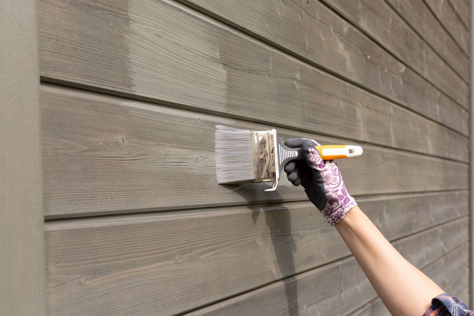 Exterior wooden siding being coated with fresh paint using a handheld brush.