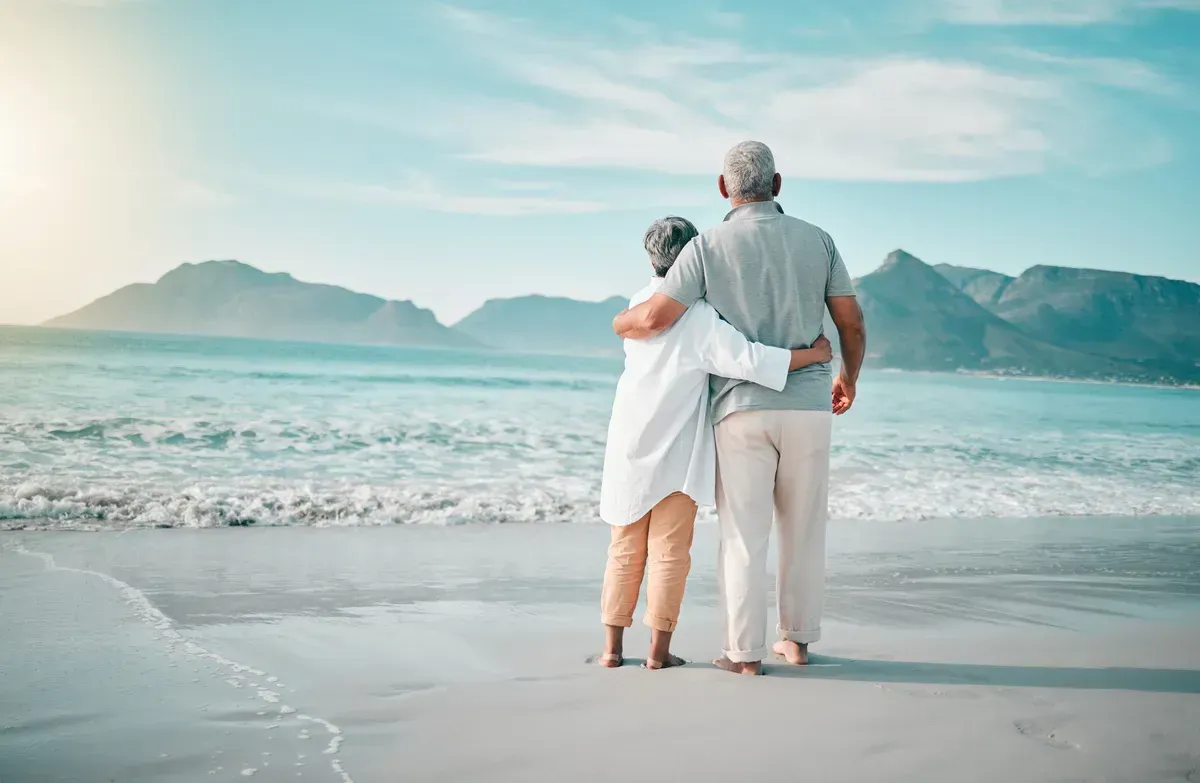 Couple standing arm in arm on a sandy beach, facing calm turquoise water and distant mountains.