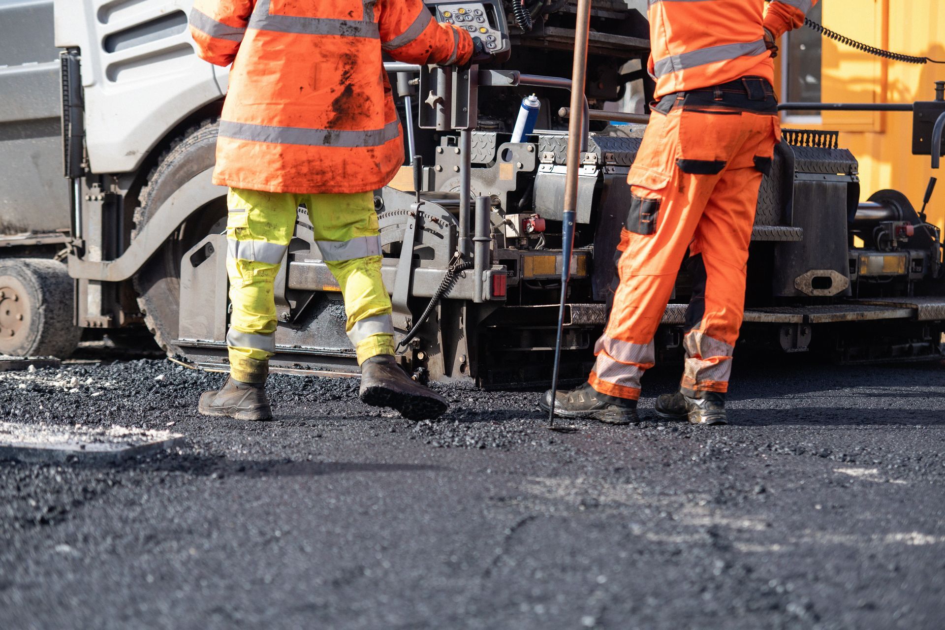 Road workers in orange safety gear paving asphalt beside a truck and paving machine Road workers in orange safety gear paving asphalt beside a truck and paving machine
