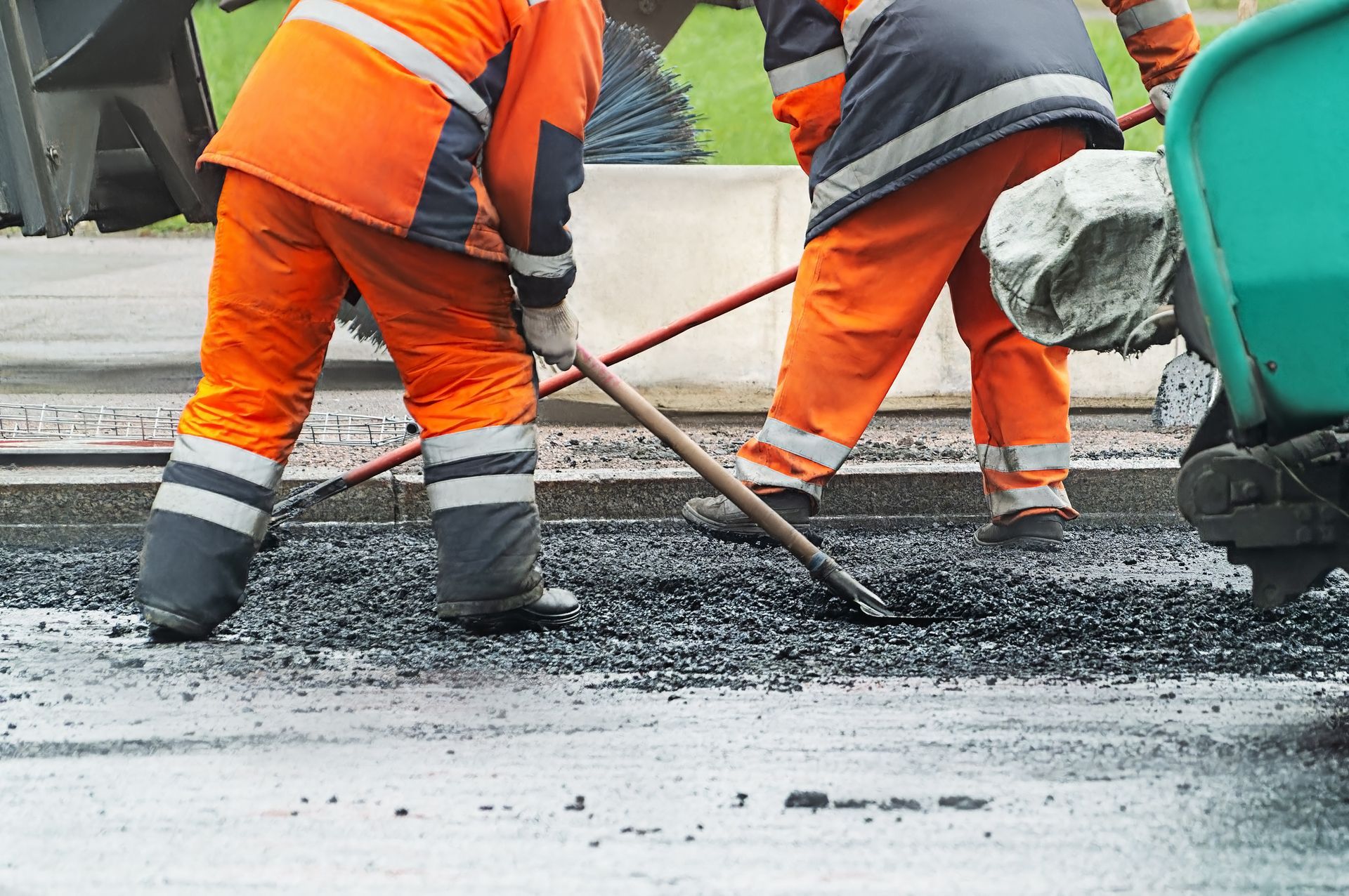 Two men in orange work clothes are shoveling asphalt.